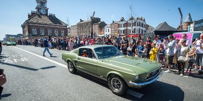 An old Ford Mustang, green in colour, travels along Stockton High Street. Crowds line the route. Stockton Town Hall is visible in the background, and you can just make out some other supercars following.