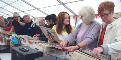 A row of people browsing through rows of records at a Record Fair - the focal point of the image is a girl with brown hair in yellow jumper holding up a Tracy Chapman record while talking to an older lady with white hair and wearing a lilac cardigan.