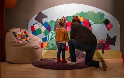 A toddler and her grown-up fill in an Elmer the Patchwork Elephant jigsaw, which is painted on a wall. A copy of the Elmer picture book rests on a chair next to them.