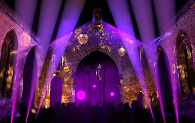 A solitary performer dangles from a wire - she is in an old church ruin, bathed in stunning purple light, and watched by a crowd.